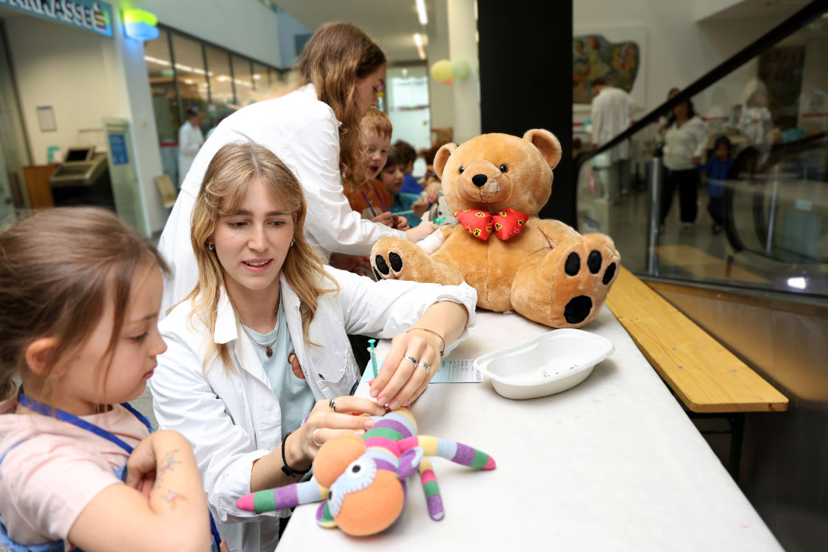 Medizinstudentin Jana Kurnik zeigt einem Kind, wie ihr Plüsch-Affe eine Spritze bekommt. Ein großer Teddy mit Pflaster sitzt daneben am Tisch im Teddybärkrankenhaus.